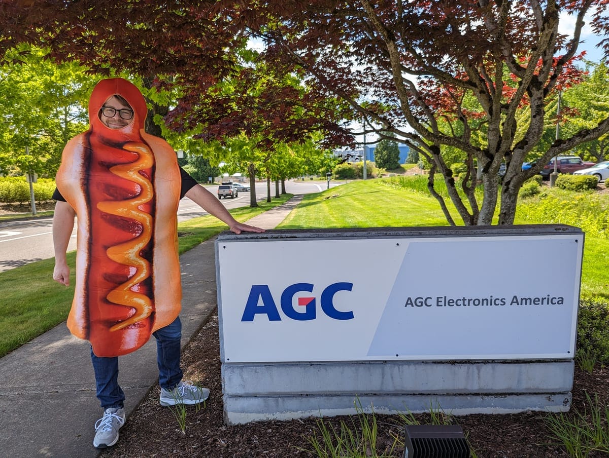 An AGCEA employee in a hot dog costume posing next to the AGC Electronics America entrance sign at the Hillsboro campus